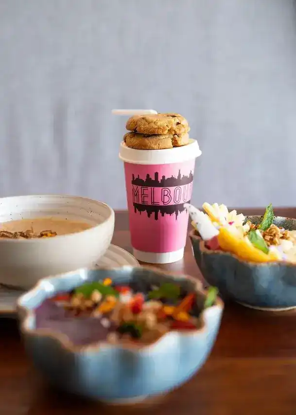 Cookies stacked on a pink Melbourne takeaway cup beside fruit and granola bowls