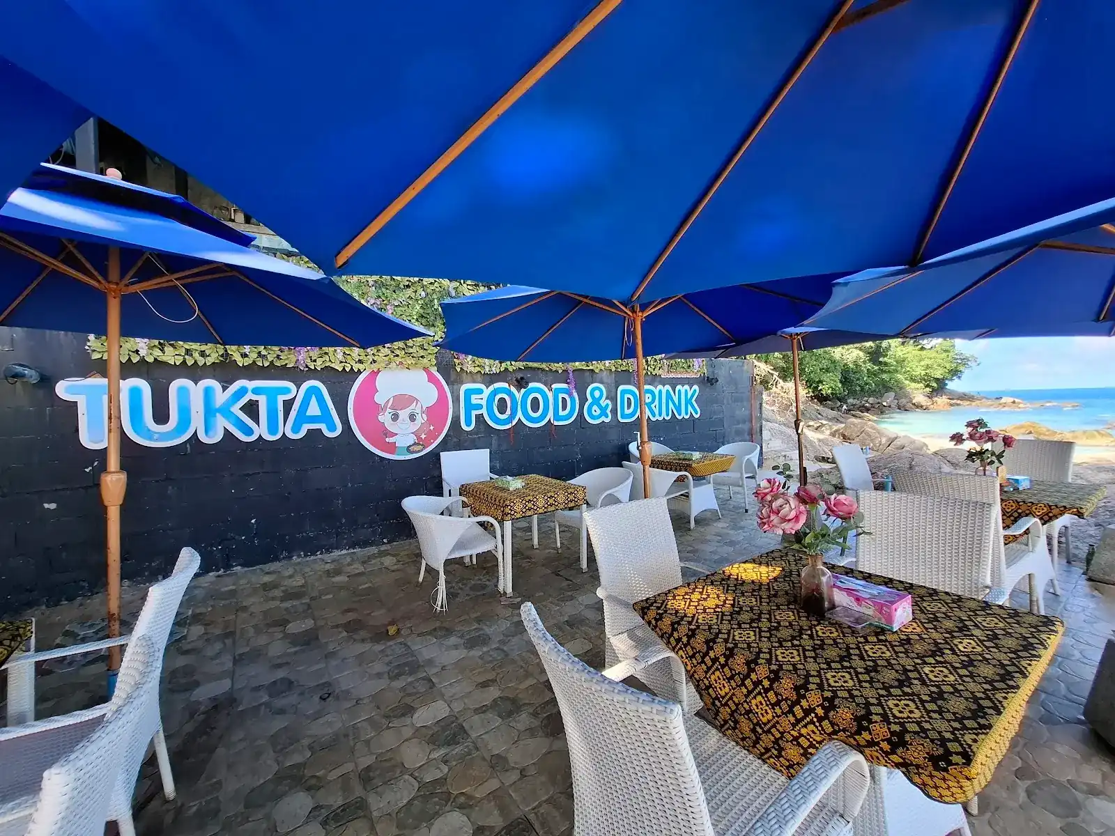Tukta's terrace: white wicker chairs and black-and-gold printed tablecloths under big blue umbrellas, the painted Tukta sign on the wall, rocks and sea on the right.