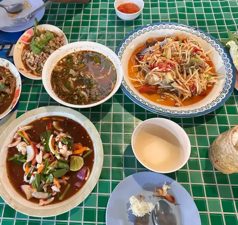 Soups, larb and a papaya salad on a green tiled tablecloth, with sticky rice in a woven basket