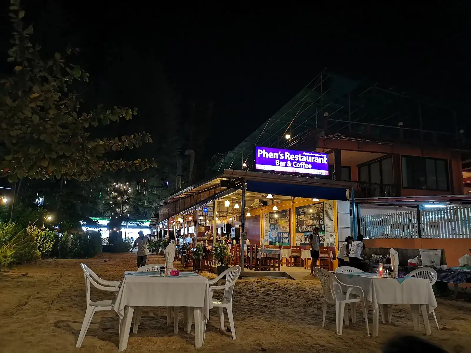 Phen's lit purple sign at night, above tables set on the sand