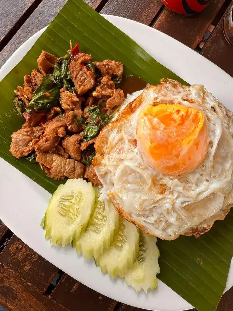 Stir-fried pork with basil and a crisp fried egg on banana leaf, with sliced cucumber