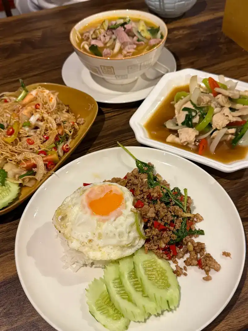 A table of Thai dishes: a bowl of soup, a spicy salad, a stir-fry, and rice with basil stir-fry and a fried egg