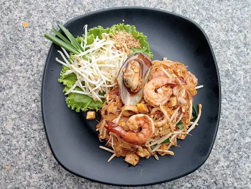 Pad thai with prawns and a mussel, bean sprouts, crushed peanuts and spring onion on a black plate.