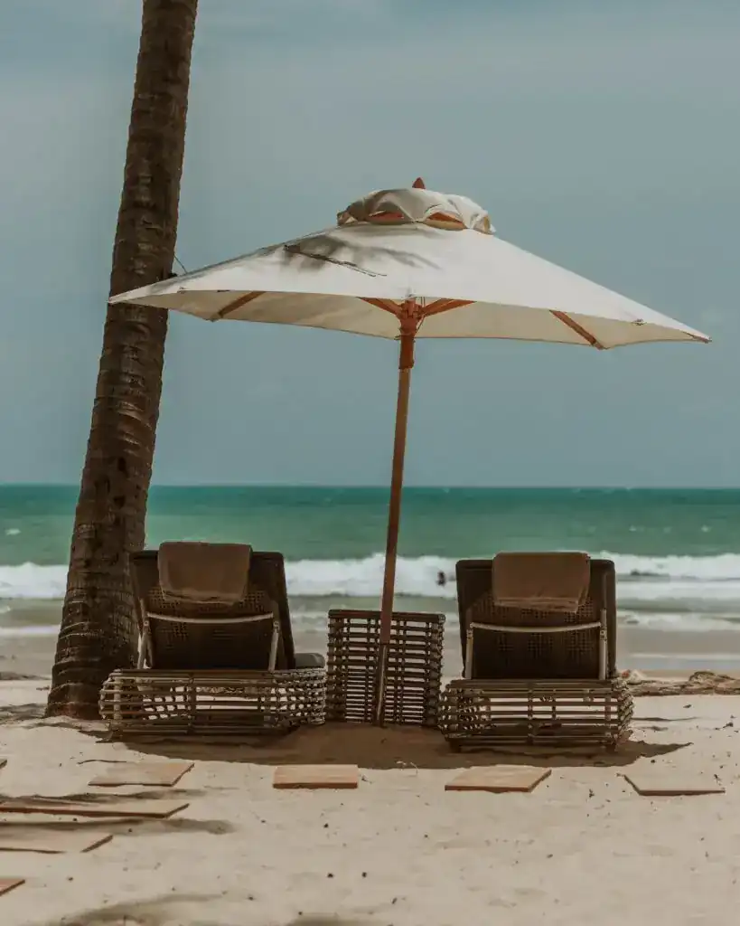 Two sun beds under a white umbrella beside a palm tree, facing the sea