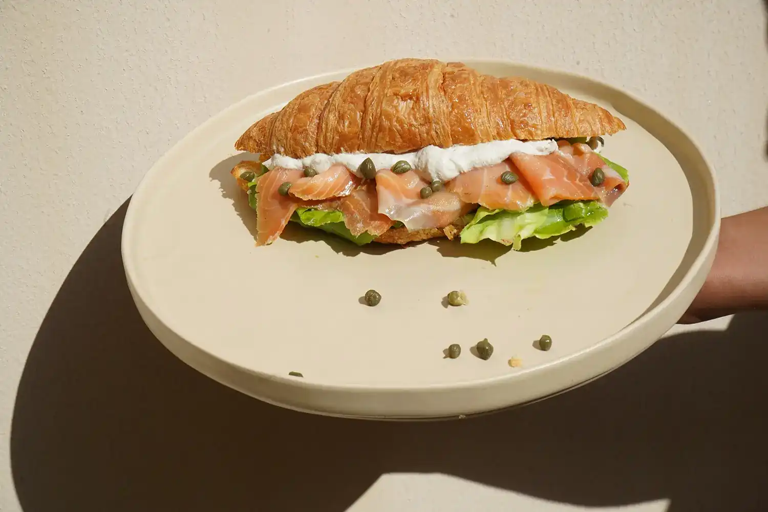 A croissant filled with smoked salmon, cream cheese, capers and lettuce on a pale plate