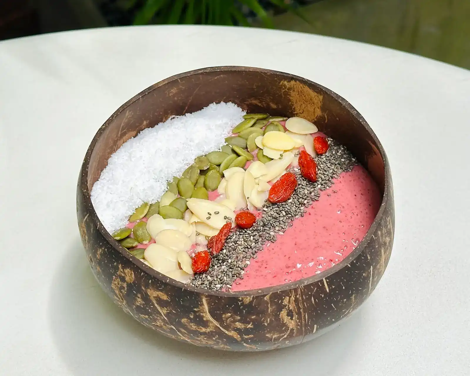 A pink smoothie bowl in a coconut shell, topped with coconut, pumpkin seeds, almonds, goji and chia