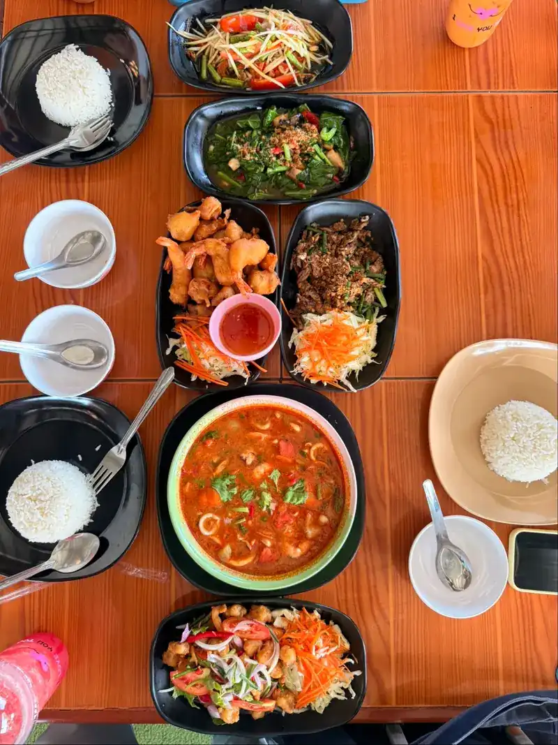 A shared table from above: rice, papaya salad, stir-fried greens, fried chicken, a spicy salad and a red soup in the middle