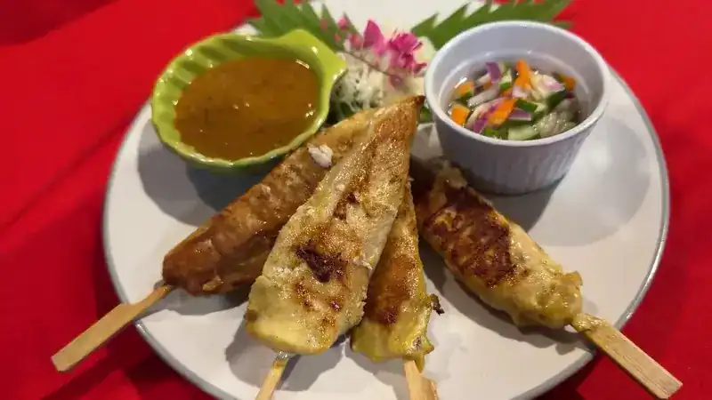 Four satay skewers with peanut sauce and cucumber relish on a red tablecloth