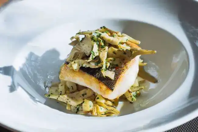 A fillet of fish with crisp skin on artichokes, in a white bowl