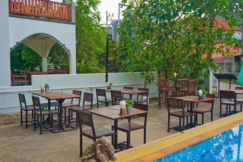 Tables under the trees on the terrace, at the yellow-tiled edge of the pool