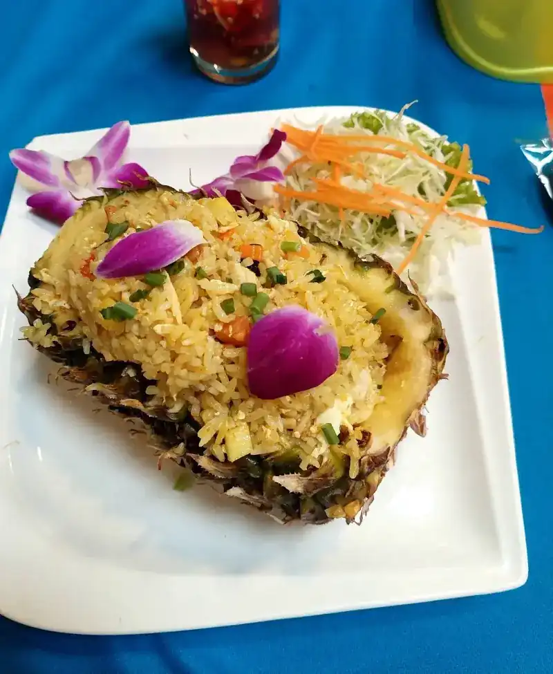 Fried rice served in half a pineapple with orchid petals and salad, on a blue tablecloth