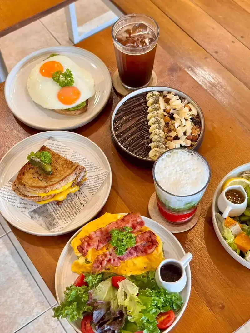 A table of breakfast plates: fried eggs on toast, a toasted sandwich, scrambled eggs with bacon and salad, and iced drinks