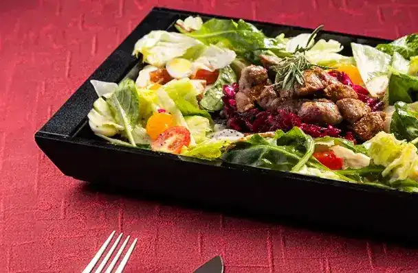 A salad topped with grilled beef in a black tray on a red velvet banquette