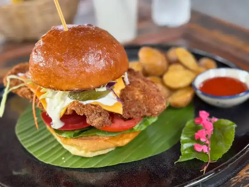 A crispy fried burger with cheese, tomato and slaw on a banana leaf, potatoes and chilli sauce behind