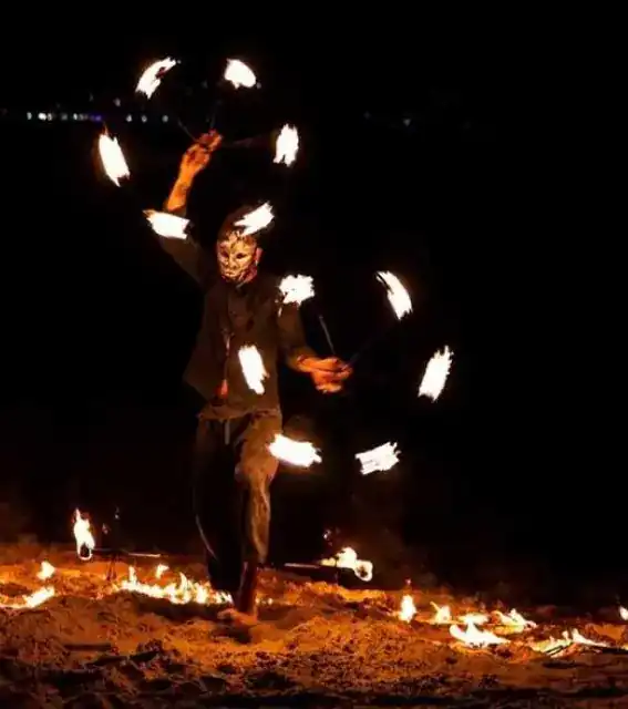 A masked fire-show performer spinning flames on the sand at night