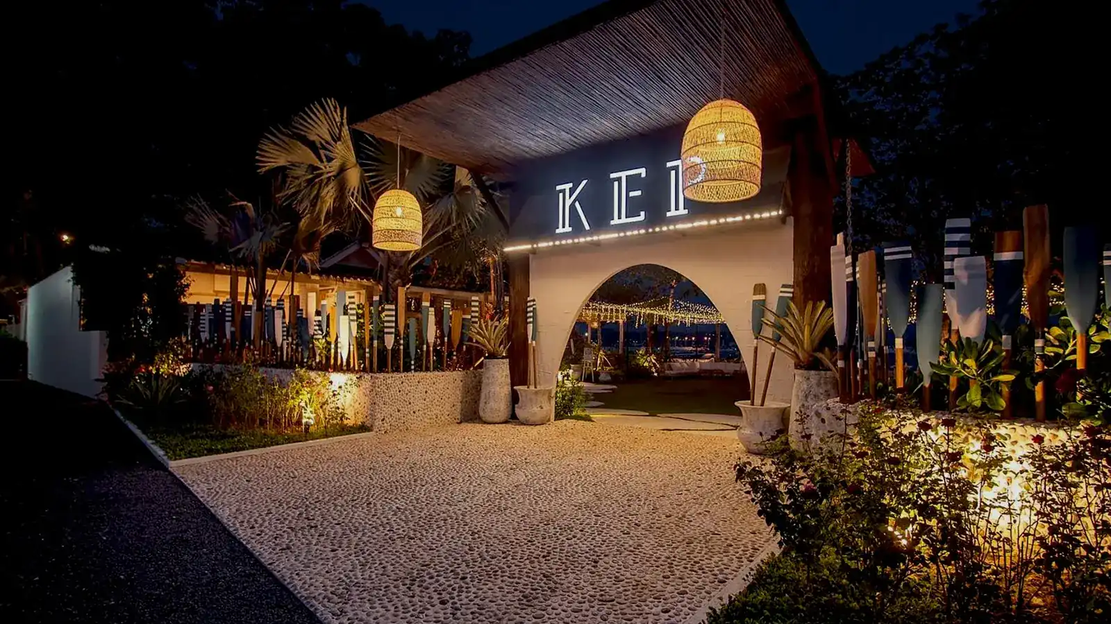 The entrance to KEP at night: a white gate with the lit KEP sign, rattan lanterns and a row of painted oars