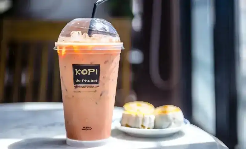 An iced drink in a Kopi de Phuket cup beside two small Phuket pastries