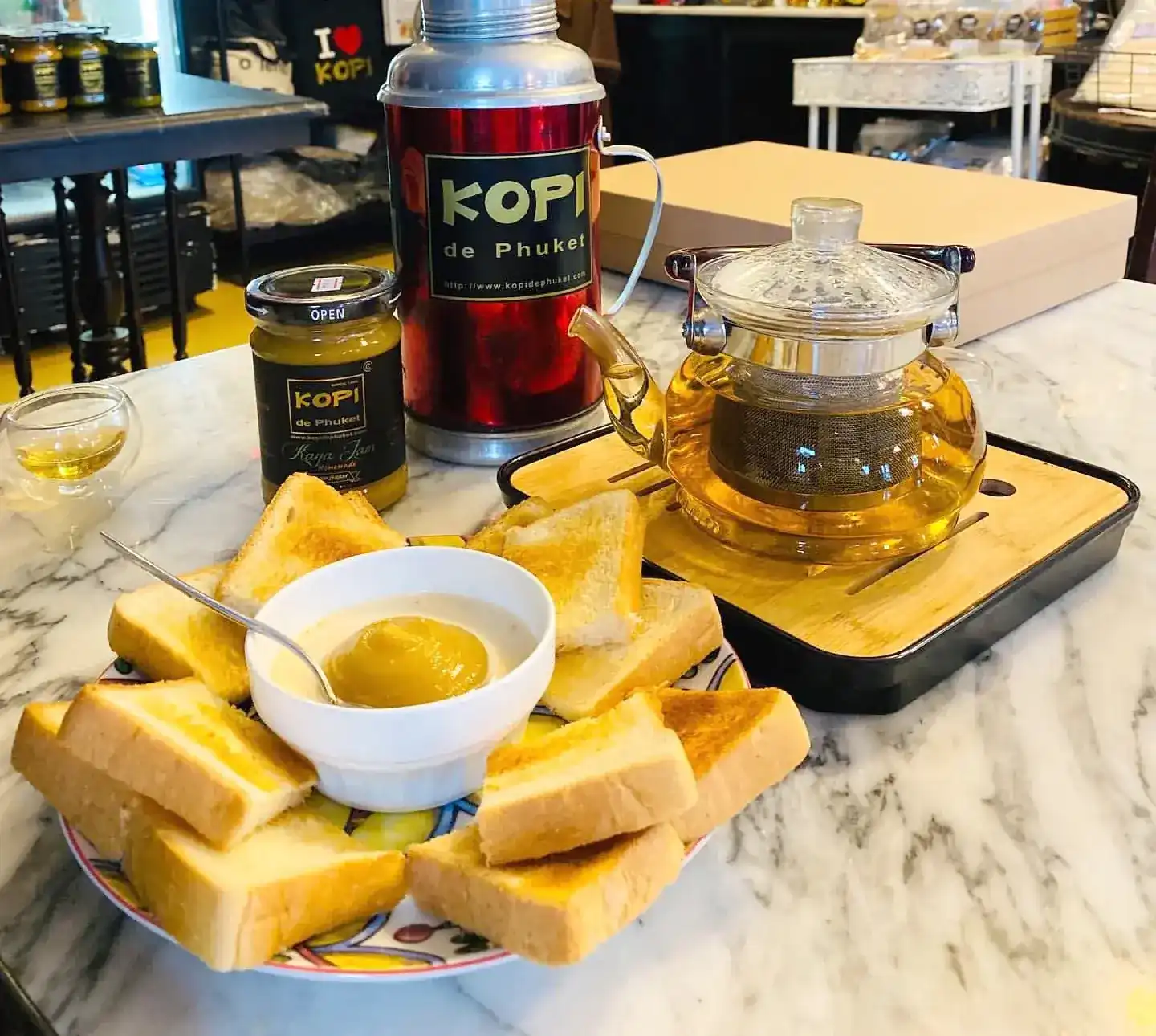 Toast with a bowl of egg kaya, a glass teapot and a red Kopi de Phuket thermos on a marble table