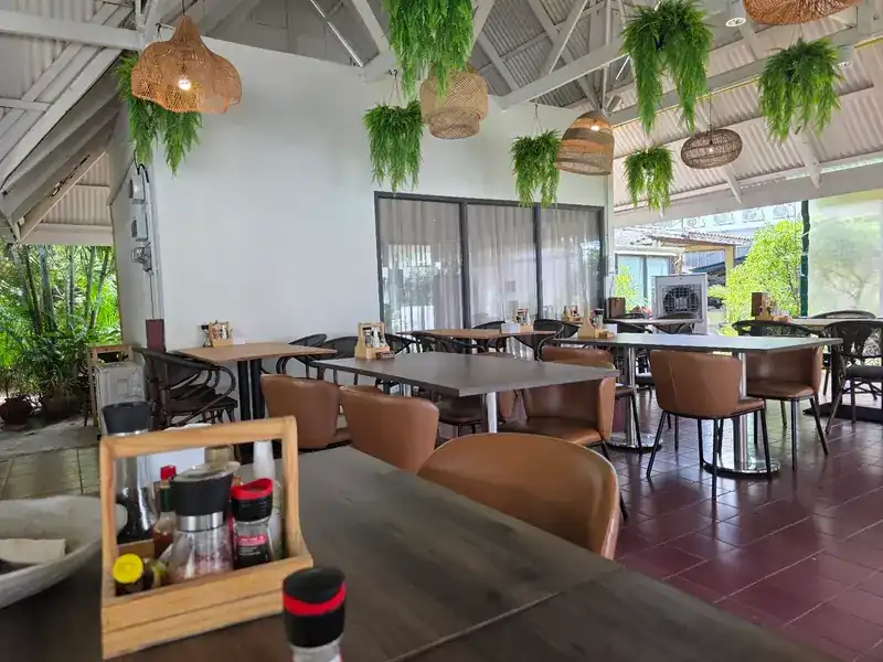 The dining room under a high roof, with hanging ferns and rattan lamps, tan leather chairs at wooden tables and greenery outside
