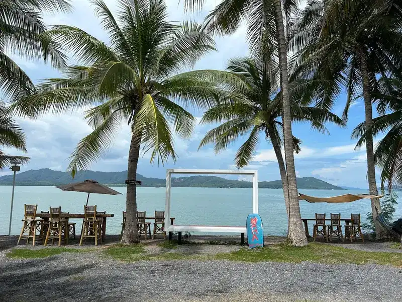 The white frame on the shore between coconut palms, with tables and bamboo chairs on either side and hills across the water