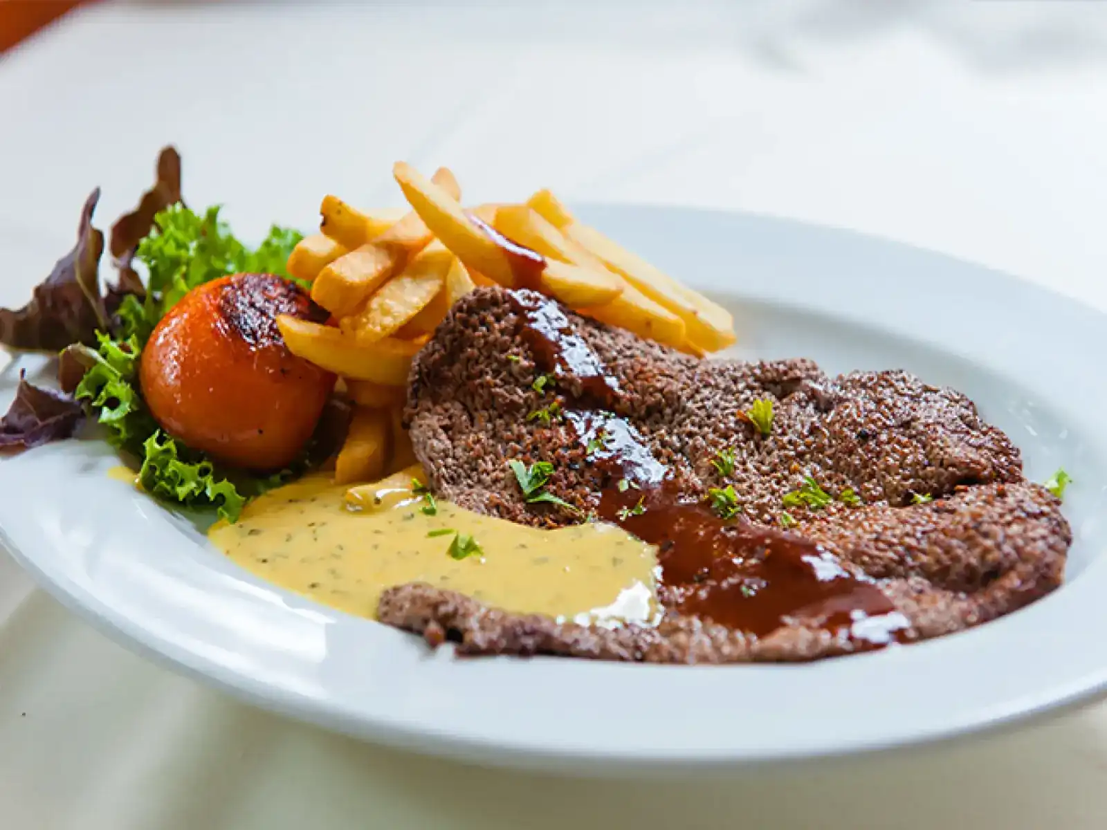 A steak on a white plate with two sauces, chips, a grilled tomato and salad leaves