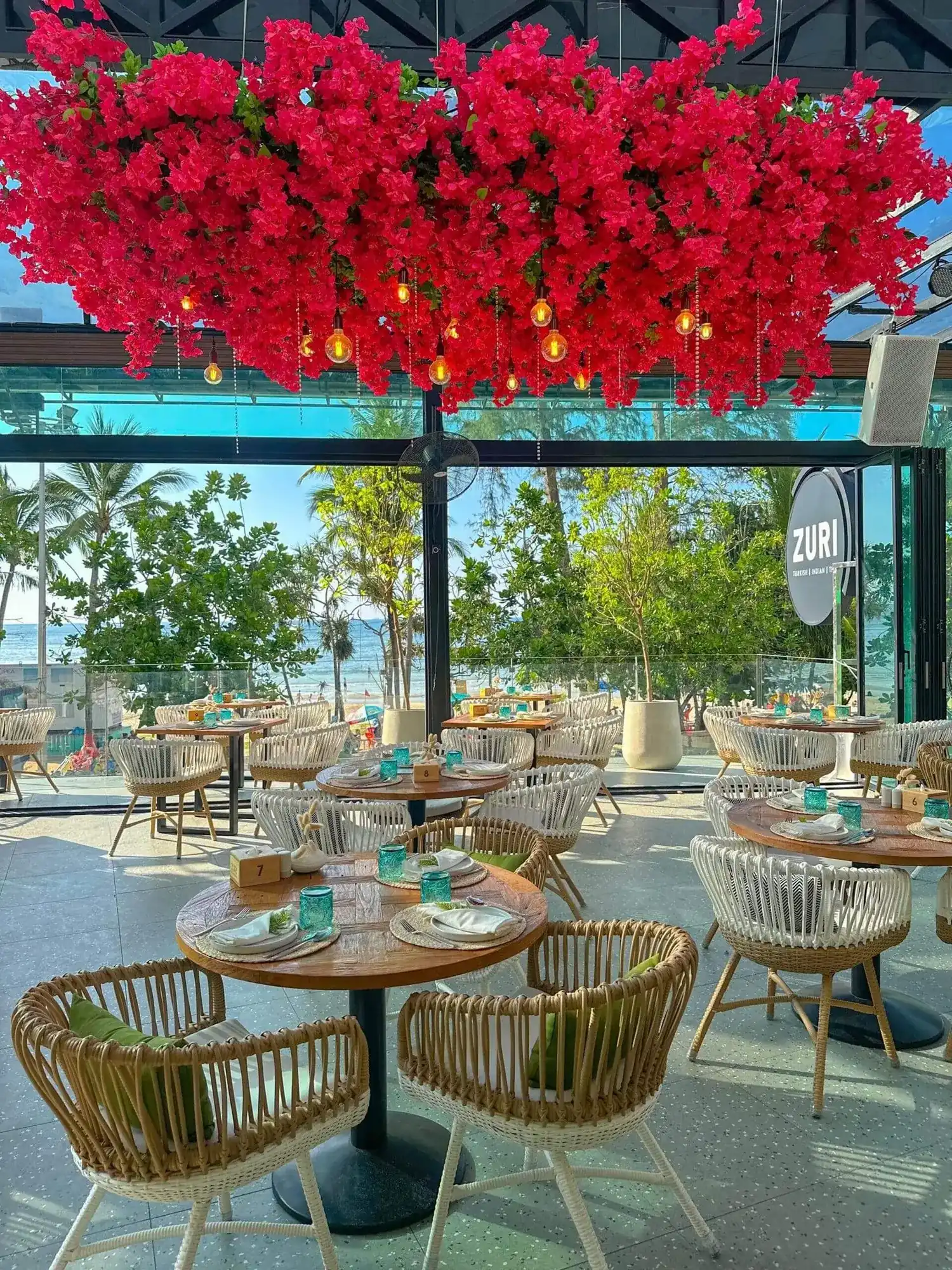 Zuri's glass terrace: rattan chairs and round tables under a canopy of red bougainvillea, with the sea at Patong Beach behind the trees