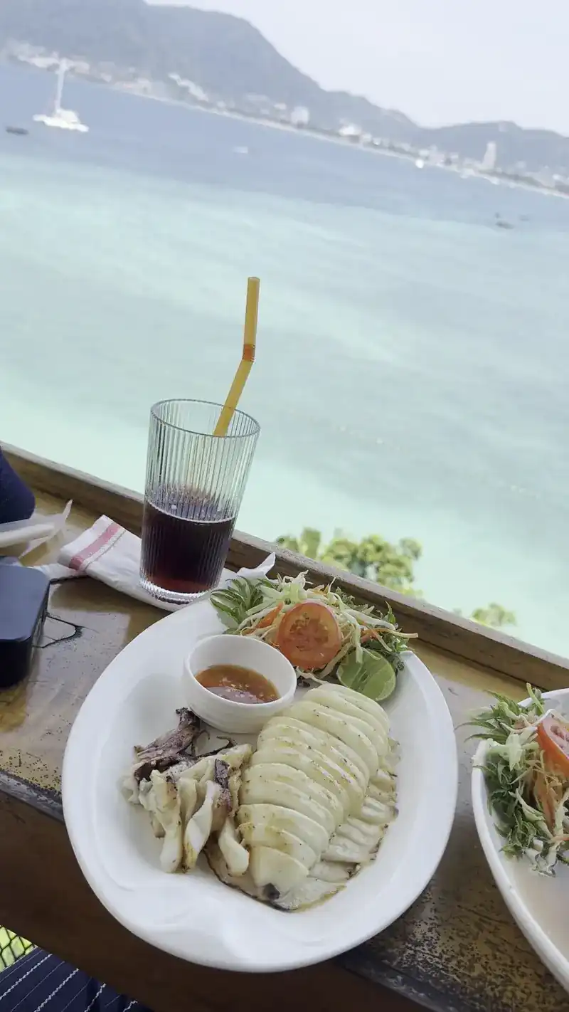 Grilled squid with salad and dipping sauce beside an iced drink, the bay and a sailing boat behind