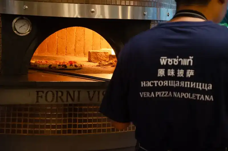 A pizza maker, seen from behind, at the mouth of the oven; his shirt reads Vera Pizza Napoletana in four languages