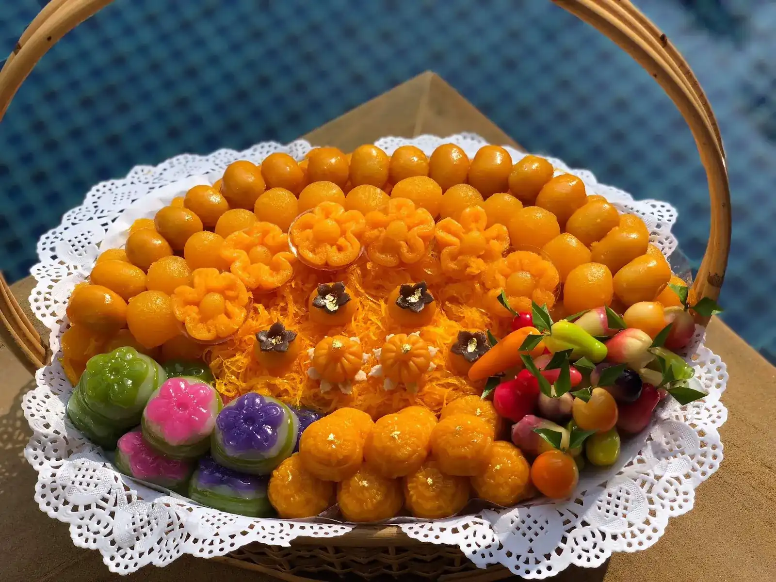 A basket of Thai sweets: golden egg-yolk sweets, flower-shaped layered sweets and fruit-shaped luk chup on a lace doily