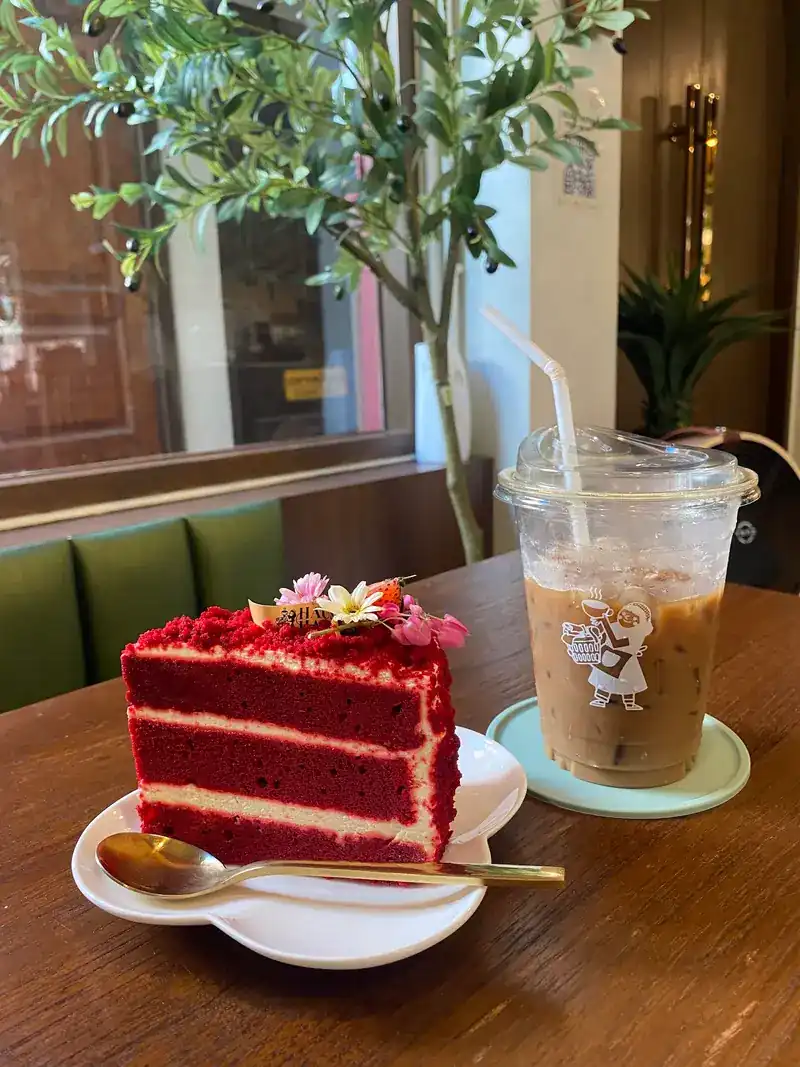 A slice of red velvet cake topped with flowers, next to an iced coffee
