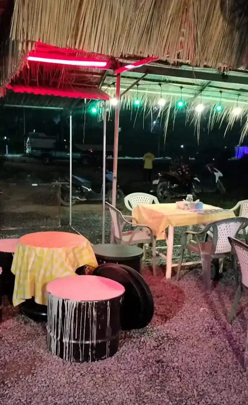 Tables with yellow checked cloths and oil-drum seats under the thatch at night, lit by red and green lights
