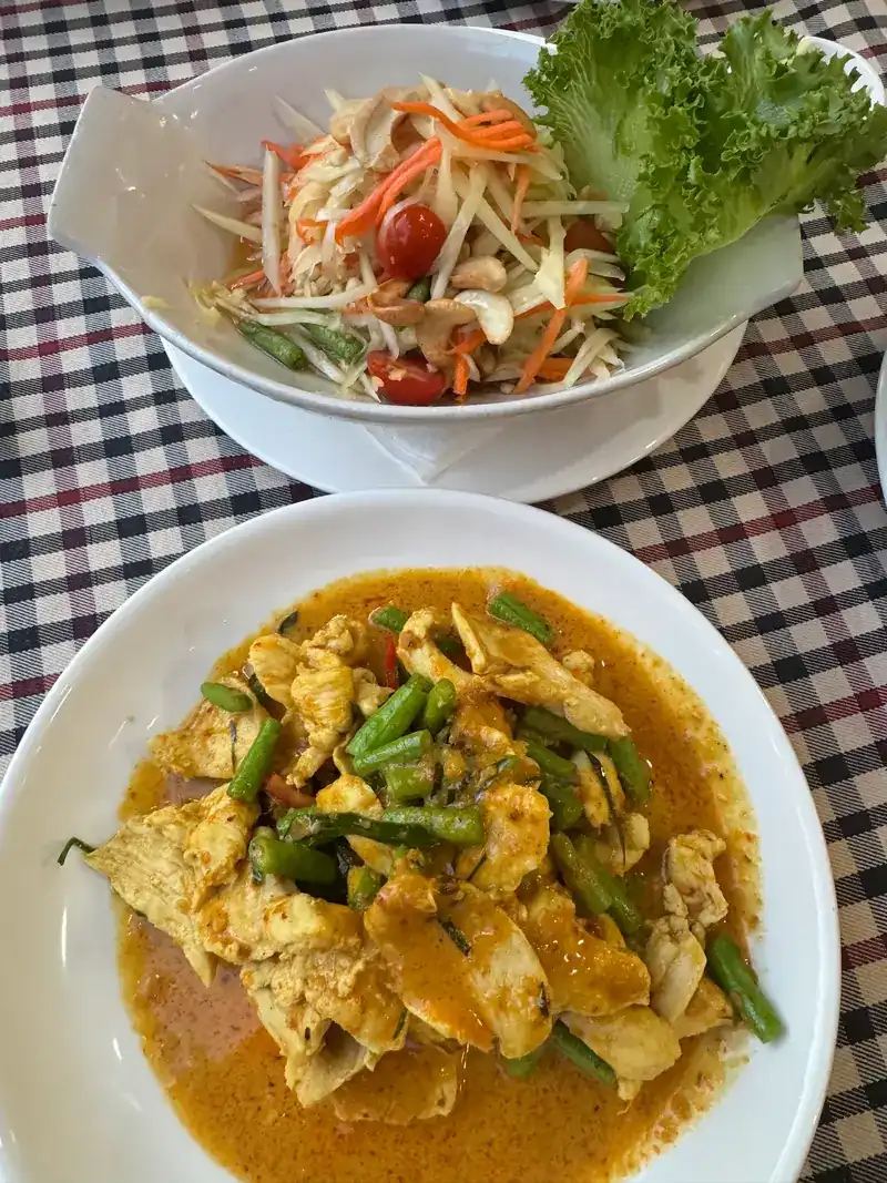A bowl of papaya salad above a plate of chicken curry with long beans, on a checked cloth