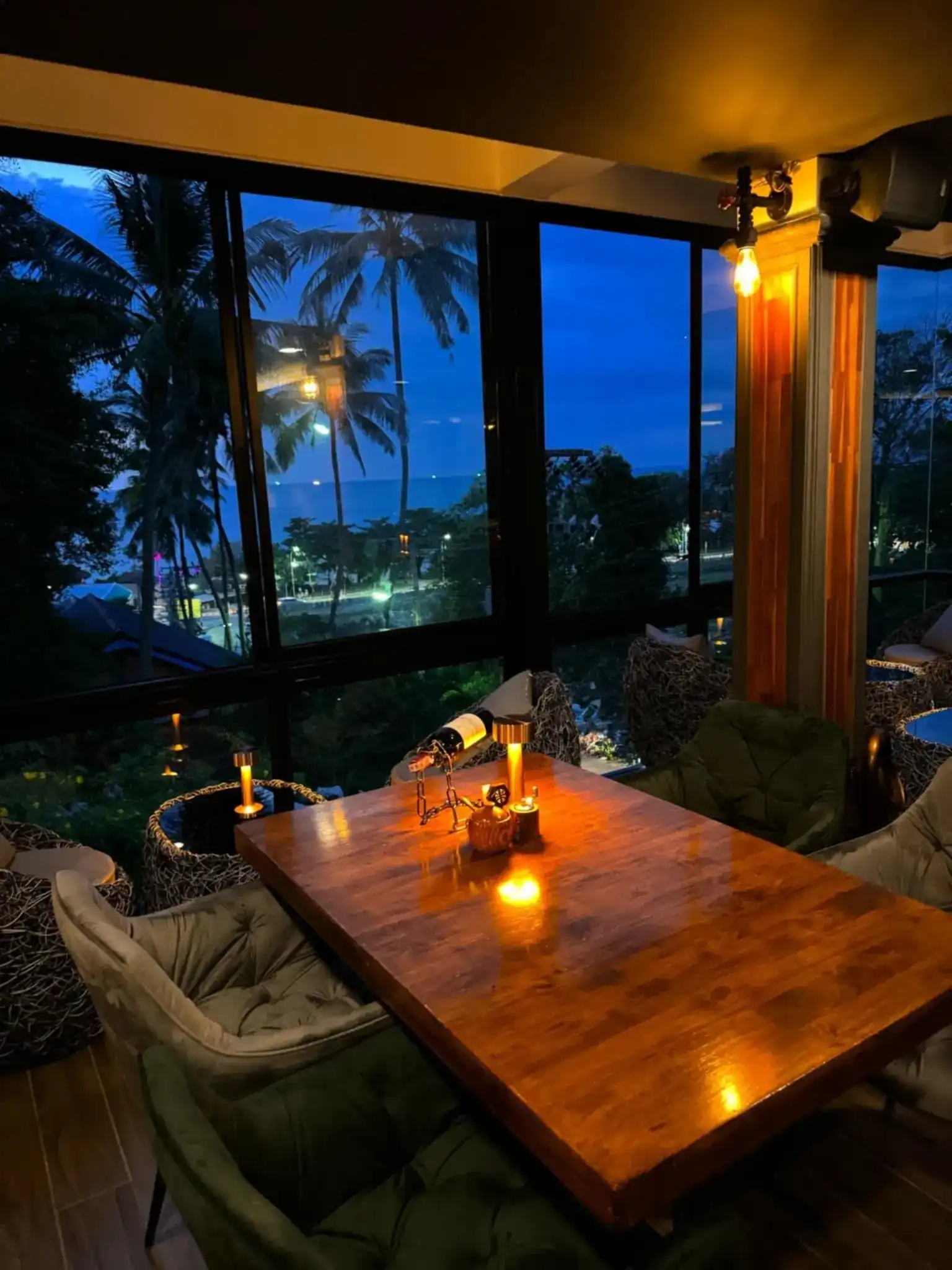 A wooden table by the windows at dusk, lit by small lamps, with palm trees and the sea outside
