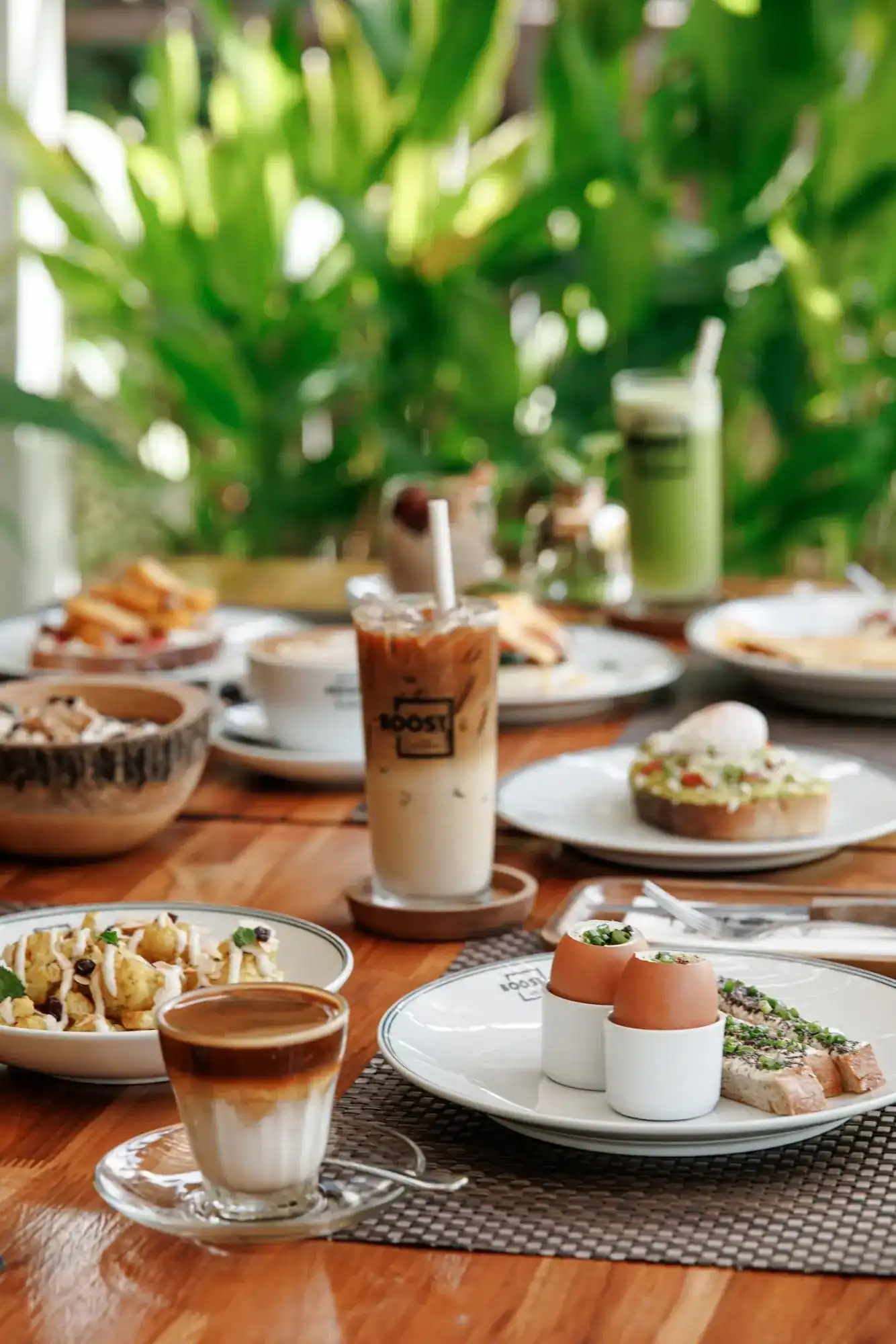 A wooden table at BOOST set with iced coffee, soft eggs, avocado toast and a layered espresso, with palms behind