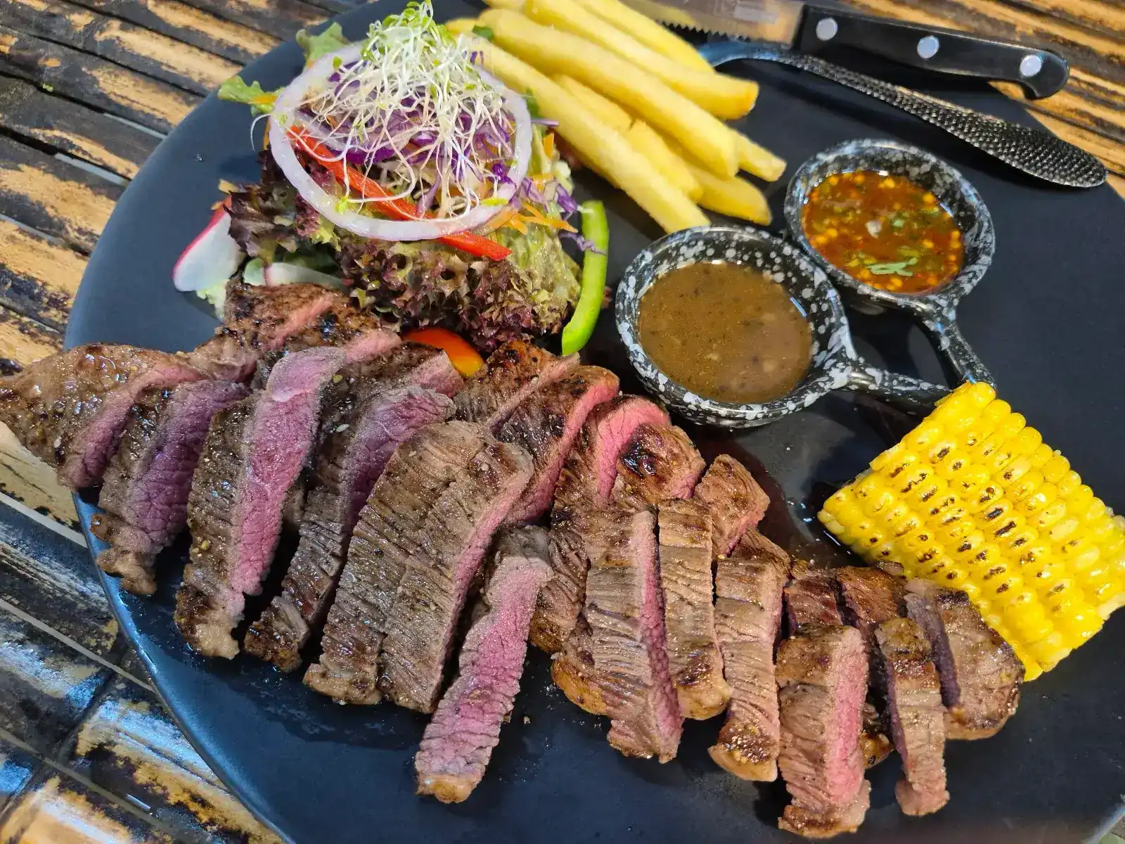 Sliced steak on a dark plate with fries, salad, grilled corn and two small pans of sauce