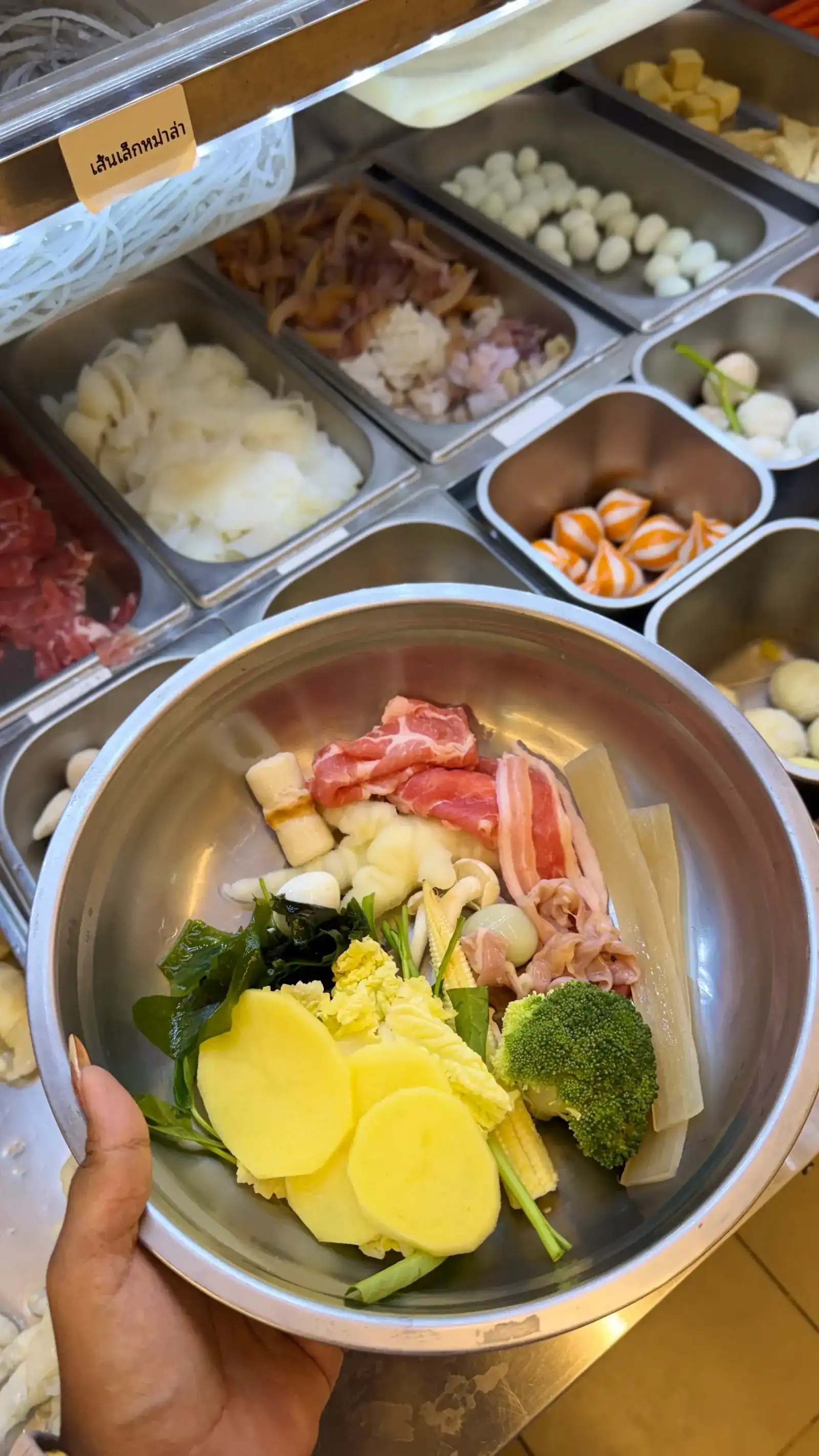 A steel bowl being filled at the chilled bar: potato, broccoli, sliced pork, baby corn and seaweed