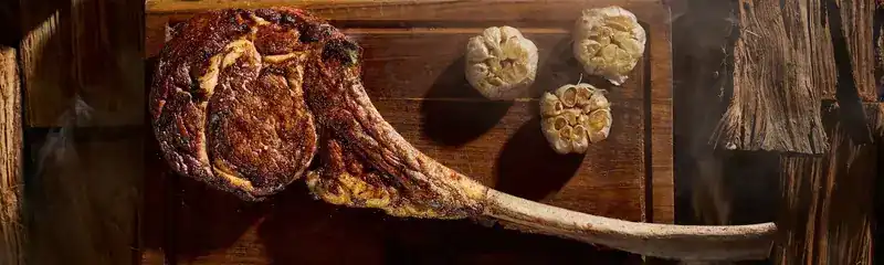 A tomahawk steak on a wooden board with three roasted garlic bulbs