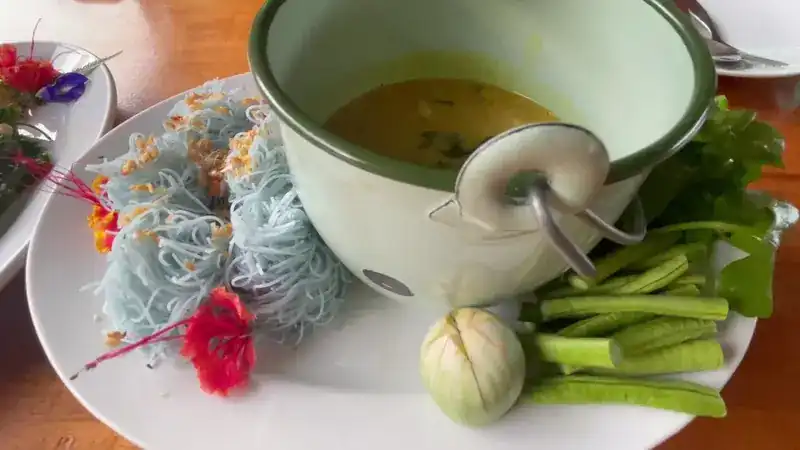Blue rice noodles with a small pot of curry and fresh vegetables on the side