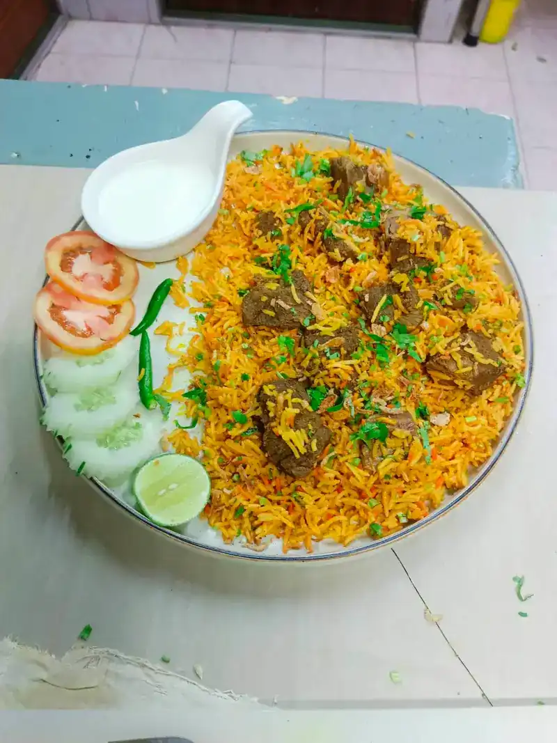 Biryani with pieces of meat, a jug of yogurt, tomato and cucumber slices, a green chilli and lime