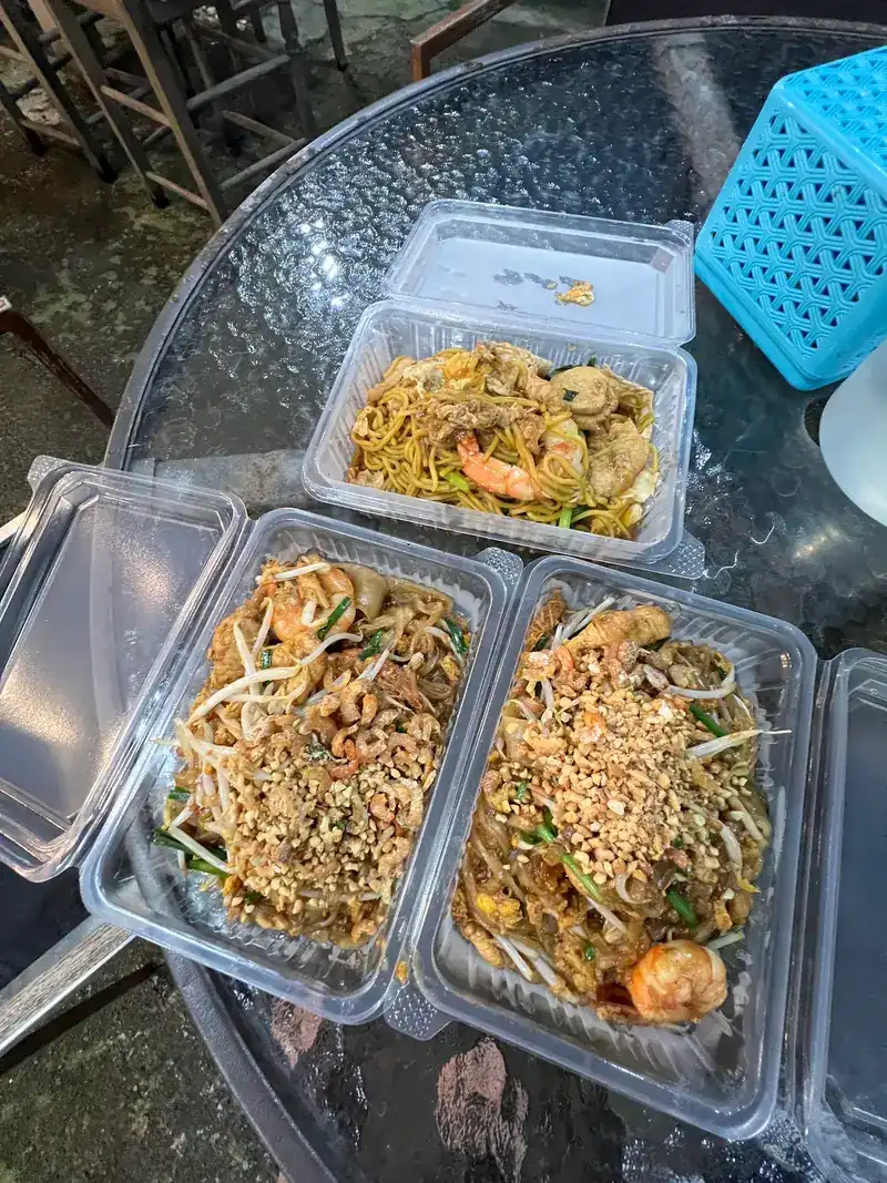 Two boxes of pad thai topped with crushed peanuts, dried shrimp and prawns, with a box of fried yellow noodles behind, on a glass table