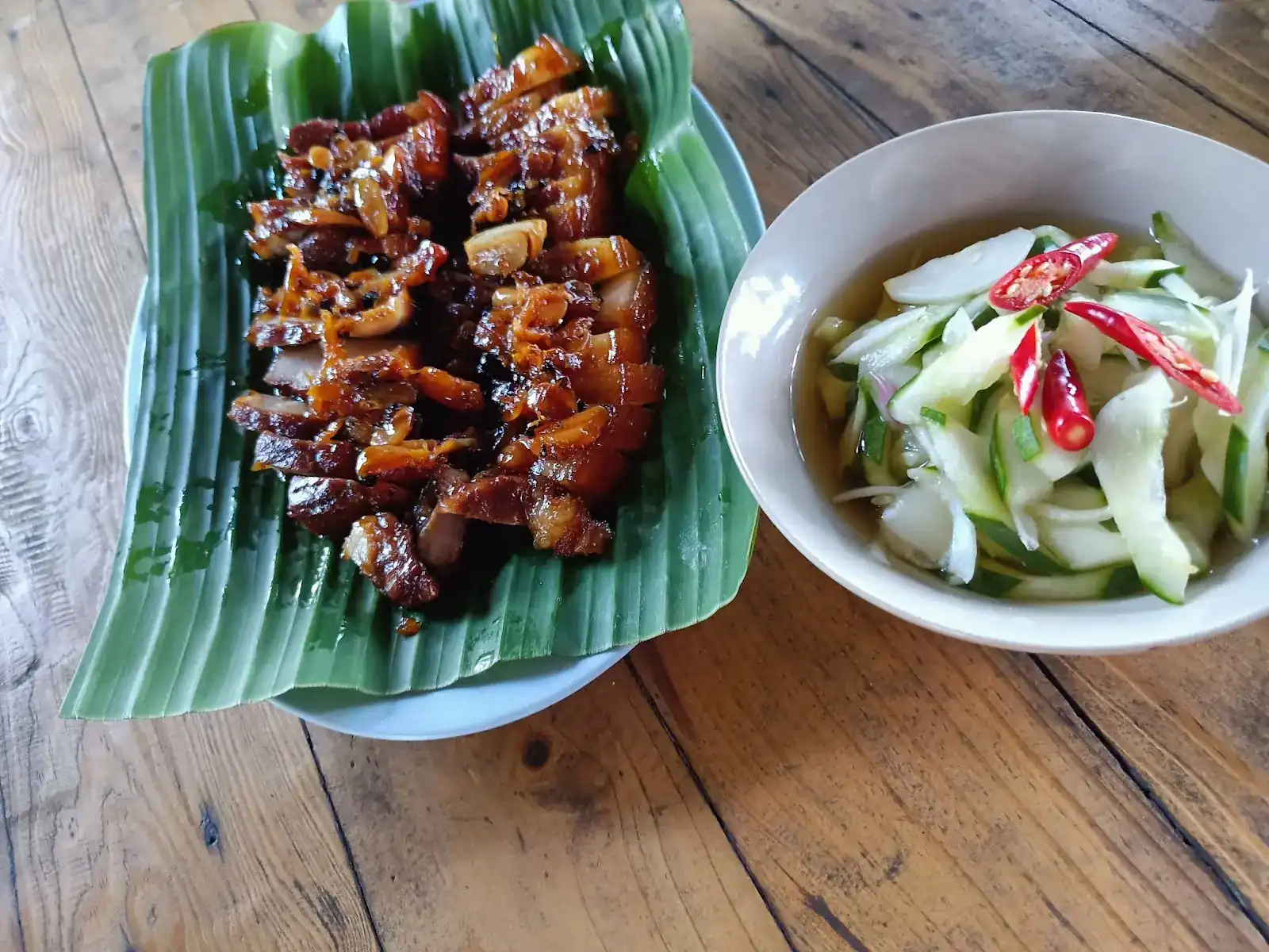 Glazed pork belly with garlic on a banana leaf, beside a bowl of cucumber and red chilli