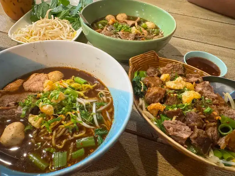 Three noodle bowls on a wooden table, one in a woven basket bowl, with bean sprouts, basil and dipping sauce