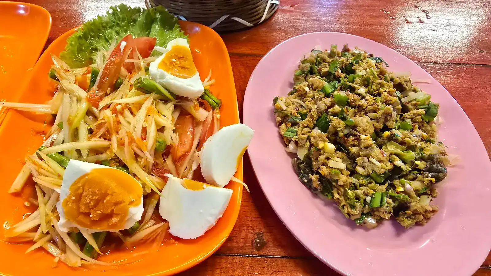 Som tam with salted egg on an orange plate, and larb on a pink plate, on a red wooden table