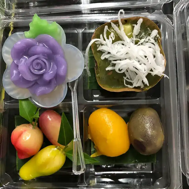 A snack box with a purple rose sweet, a coconut-topped sweet and fruit-shaped luk chup