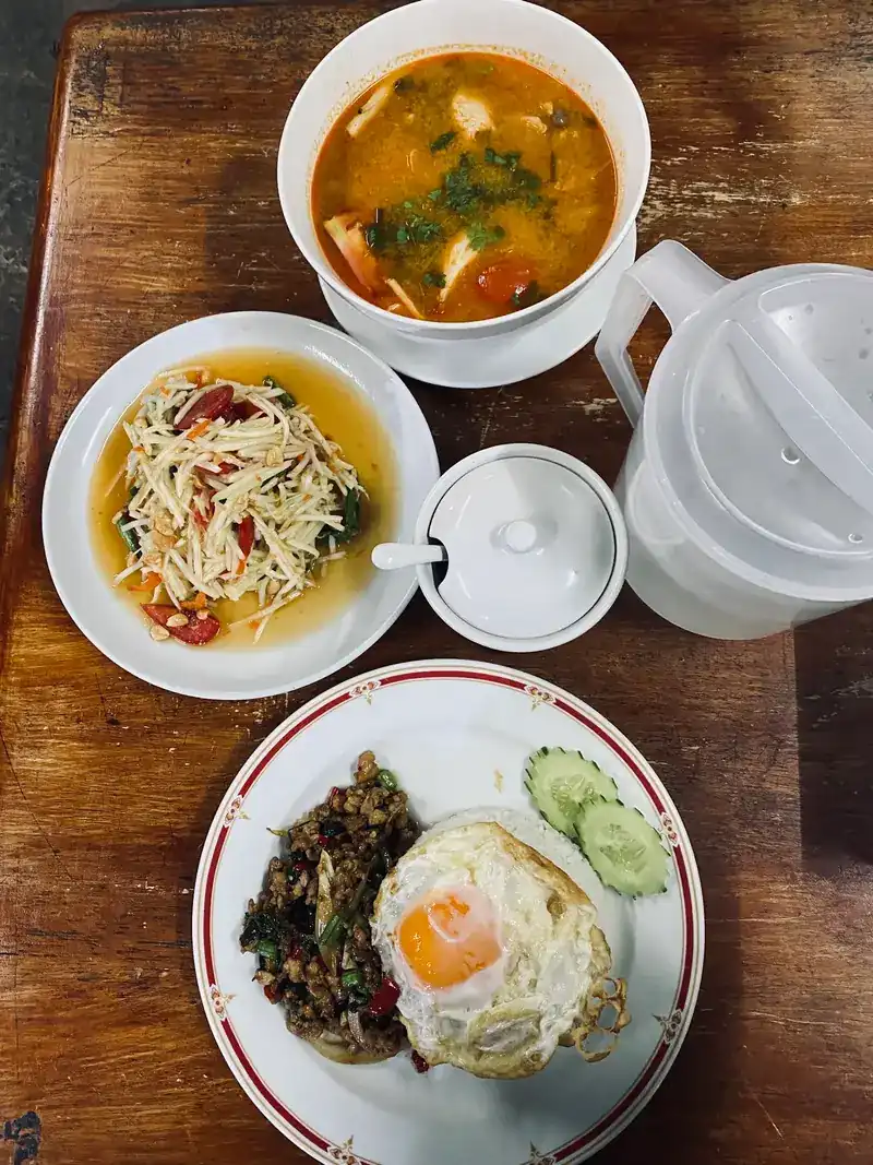 Tom yum, green papaya salad, and a basil stir-fry on rice with a fried egg, on a wooden table