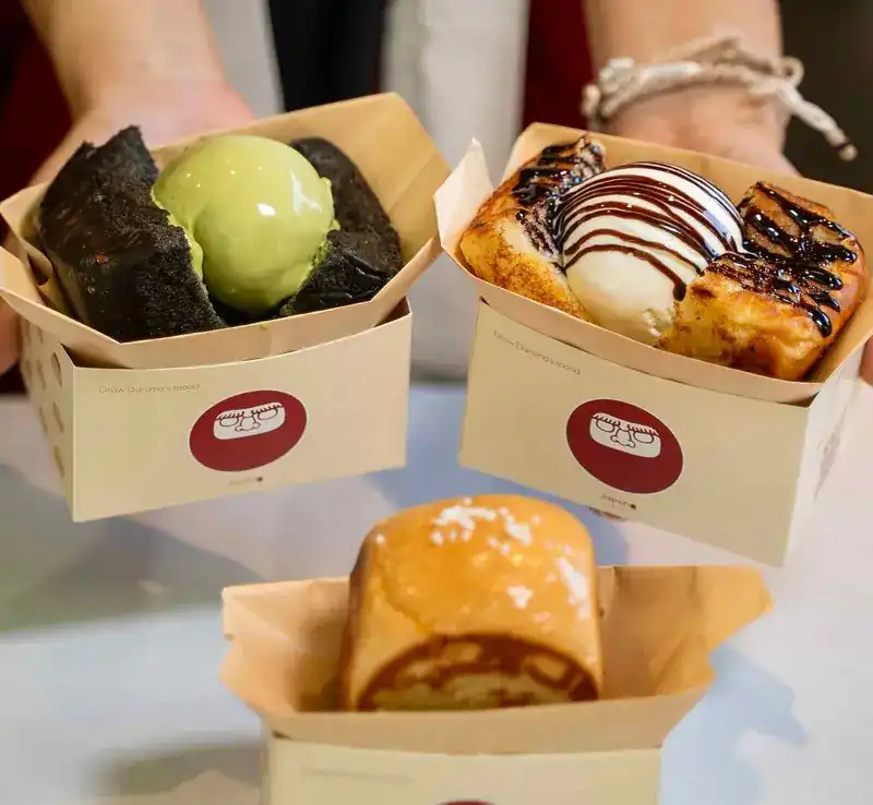 Three paper trays held out: a black bun with matcha, a golden bun with vanilla and chocolate sauce, and a plain grilled bun