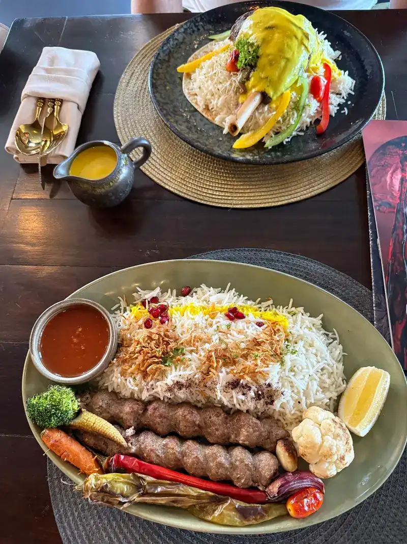 Two grilled minced-meat kebabs on white and yellow rice with grilled vegetables, lemon and a small bowl of sauce