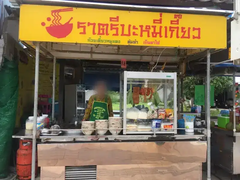 The noodle stall under the yellow Ratri Bamee Kiao sign, with stacked bowls and a glass cabinet of red pork