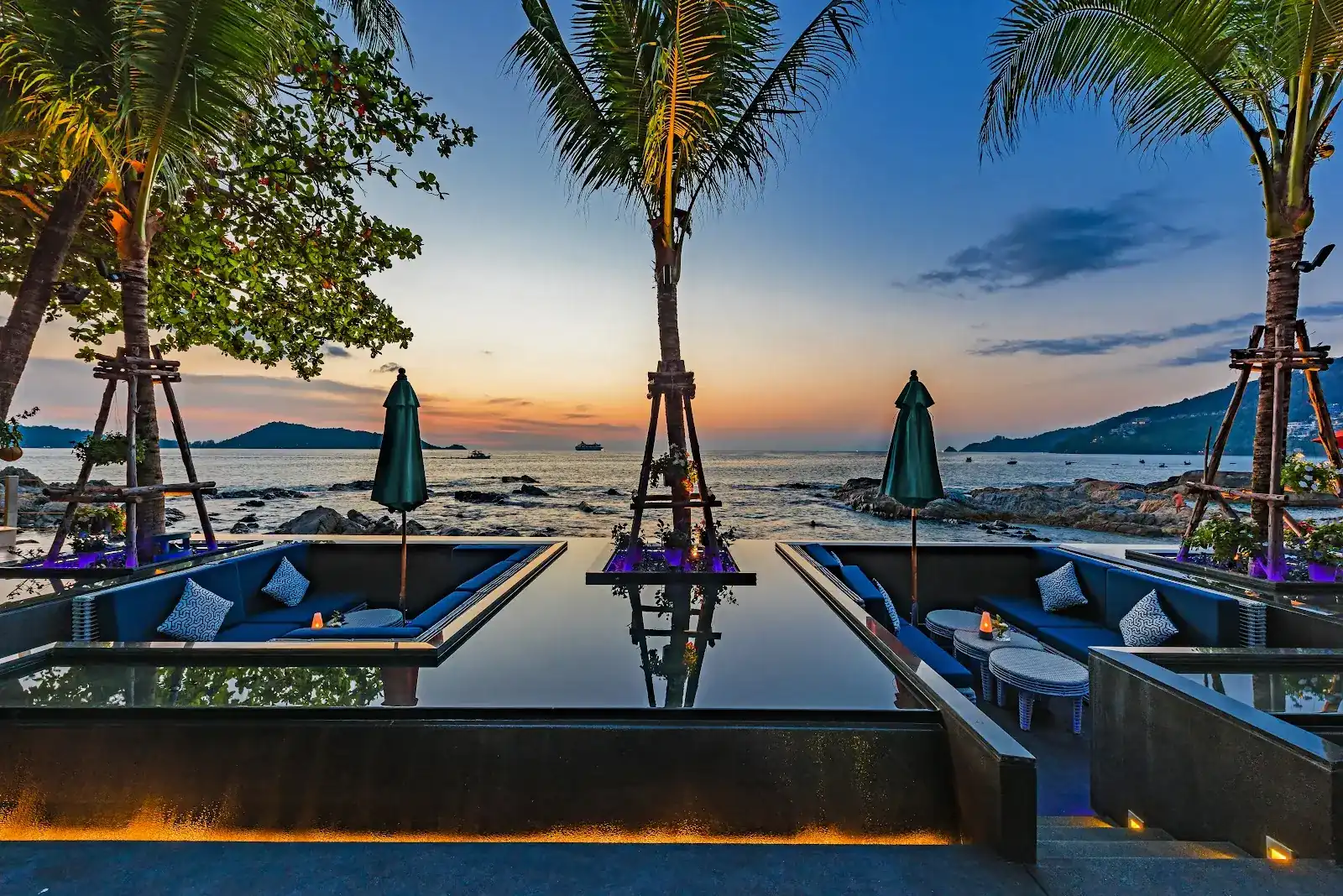 Lounge seats around a still pool at dusk, with palms and the Andaman Sea behind