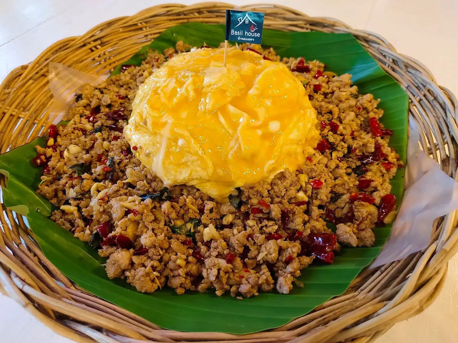 Minced pork stir-fried with holy basil and dried chillies under a soft omelette, on banana leaf in a woven basket, with a Basil House flag